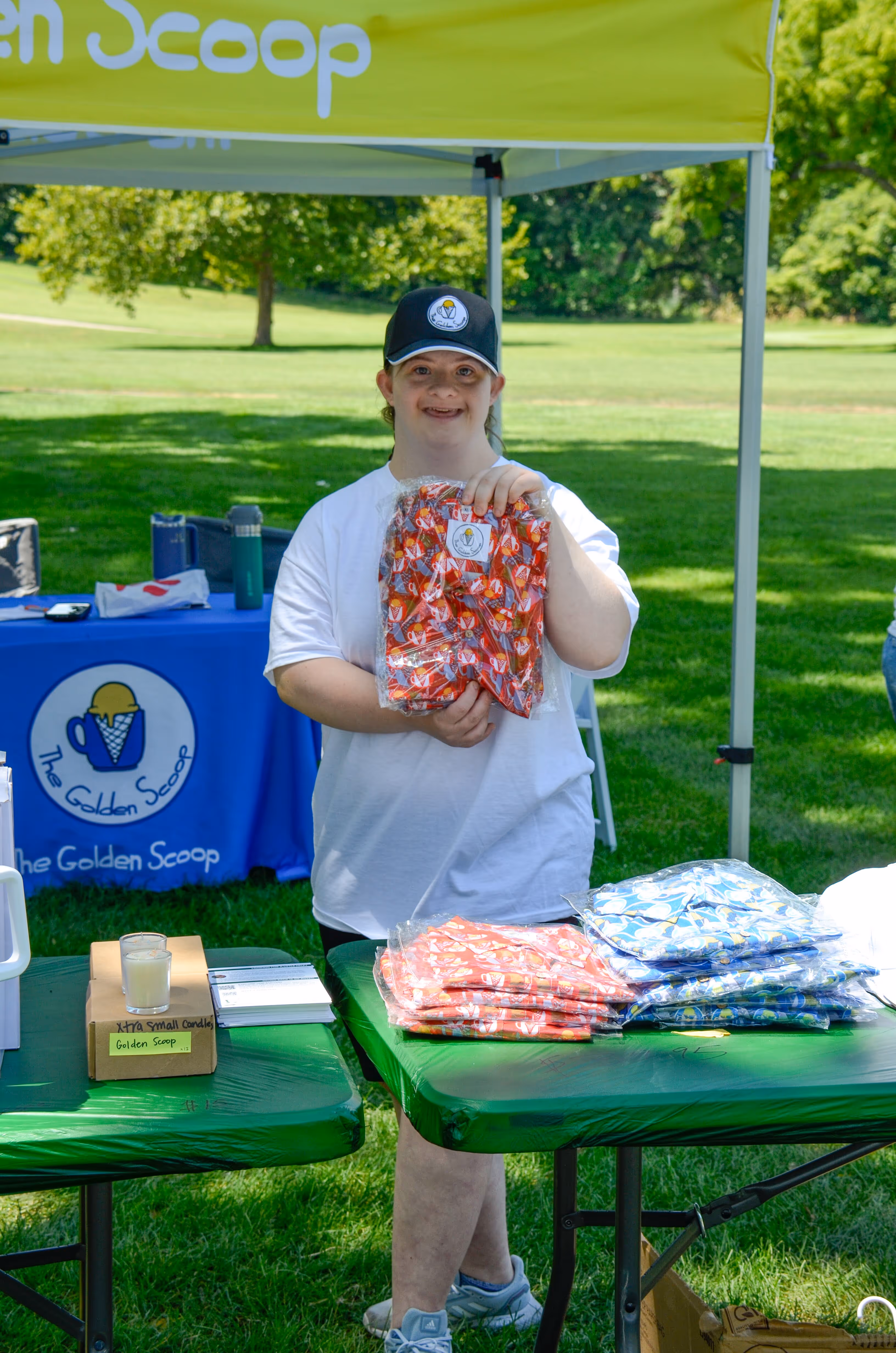 Group of people, including children and adults, gathered outdoors near a stage with some hugging and others standing nearby in casual summer clothing.