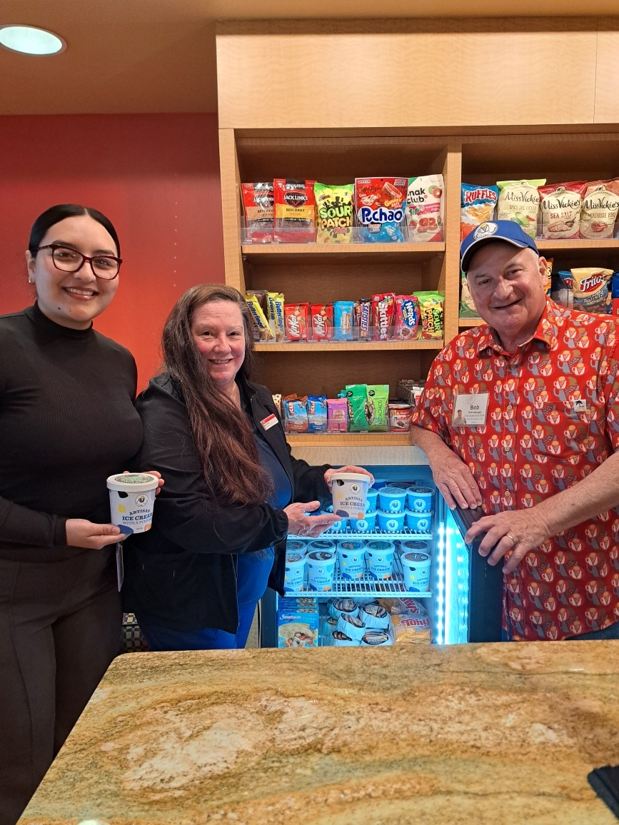 Three people smiling at a counter in front of a snack shelf and an open freezer filled with Bayfield ice cream tubs.