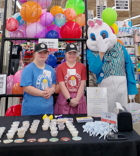 Two young women wearing ice cream-themed shirts and black caps stand behind a table with samples and spoons next to a person in a large bird mascot costume inside a store with colorful balloons behind.