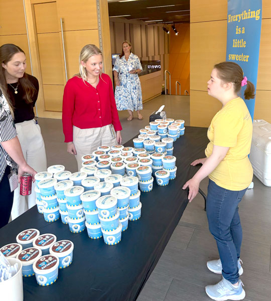 Two young women wearing ice cream-themed shirts and black caps stand behind a table with samples and spoons next to a person in a large bird mascot costume inside a store with colorful balloons behind.