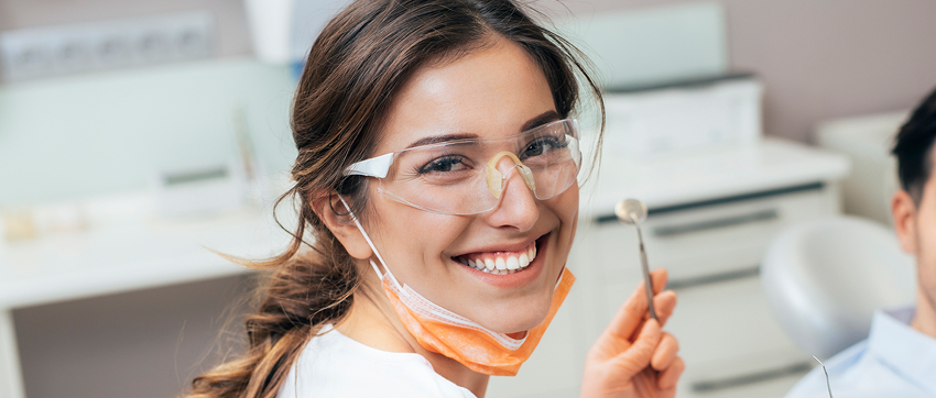 Smiling female dentist wearing protective glasses and an orange face mask, holding a dental mirror in a dental office.