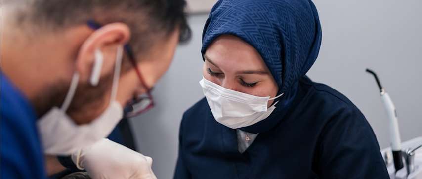 Two healthcare professionals wearing surgical masks and gloves, focused on a medical procedure, with one wearing a navy blue headscarf and uniform.