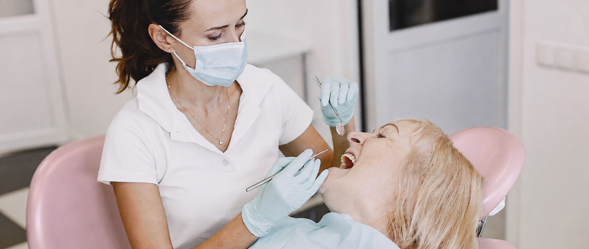 Dentist wearing a face mask and gloves examining the open mouth of a patient reclining in a dental chair.