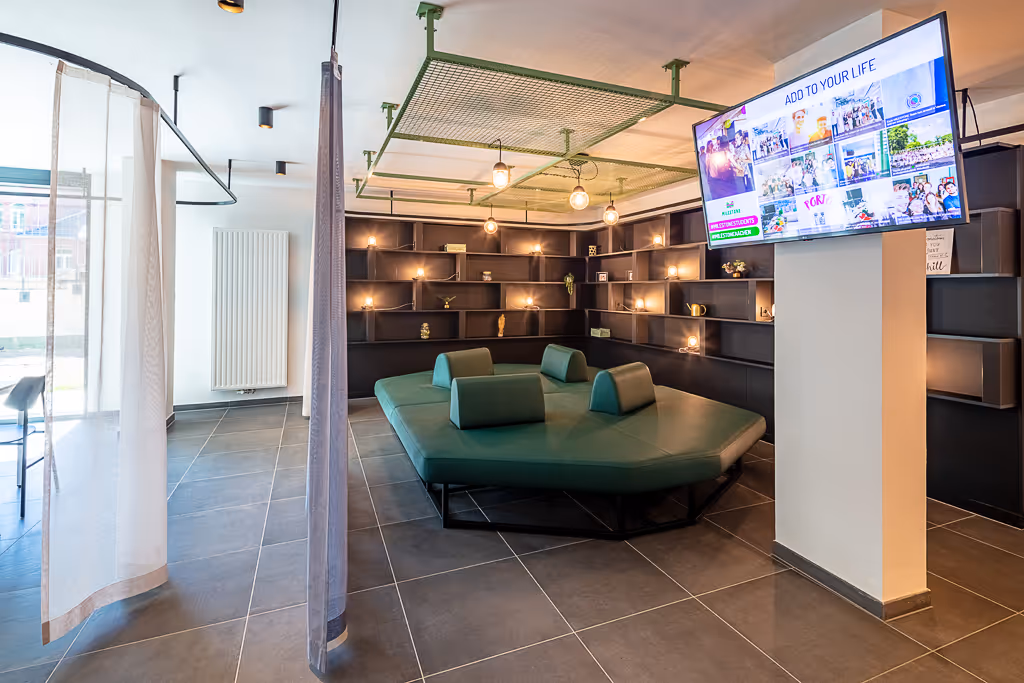 Gemütliche Gemeinschaftslounge im Studentenwohnheim „Guter Hirte“ in Aachen mit modernem grünen Polstersofa, Wandregalen, stimmungsvoller Beleuchtung und großem Bildschirm – ideal zum Entspannen und Austauschen.