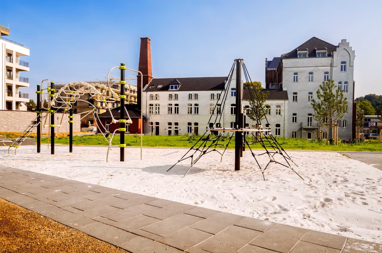Moderner Spielplatz mit Klettergerüsten und Sandfläche vor historischen Gebäuden bei klarem Himmel.