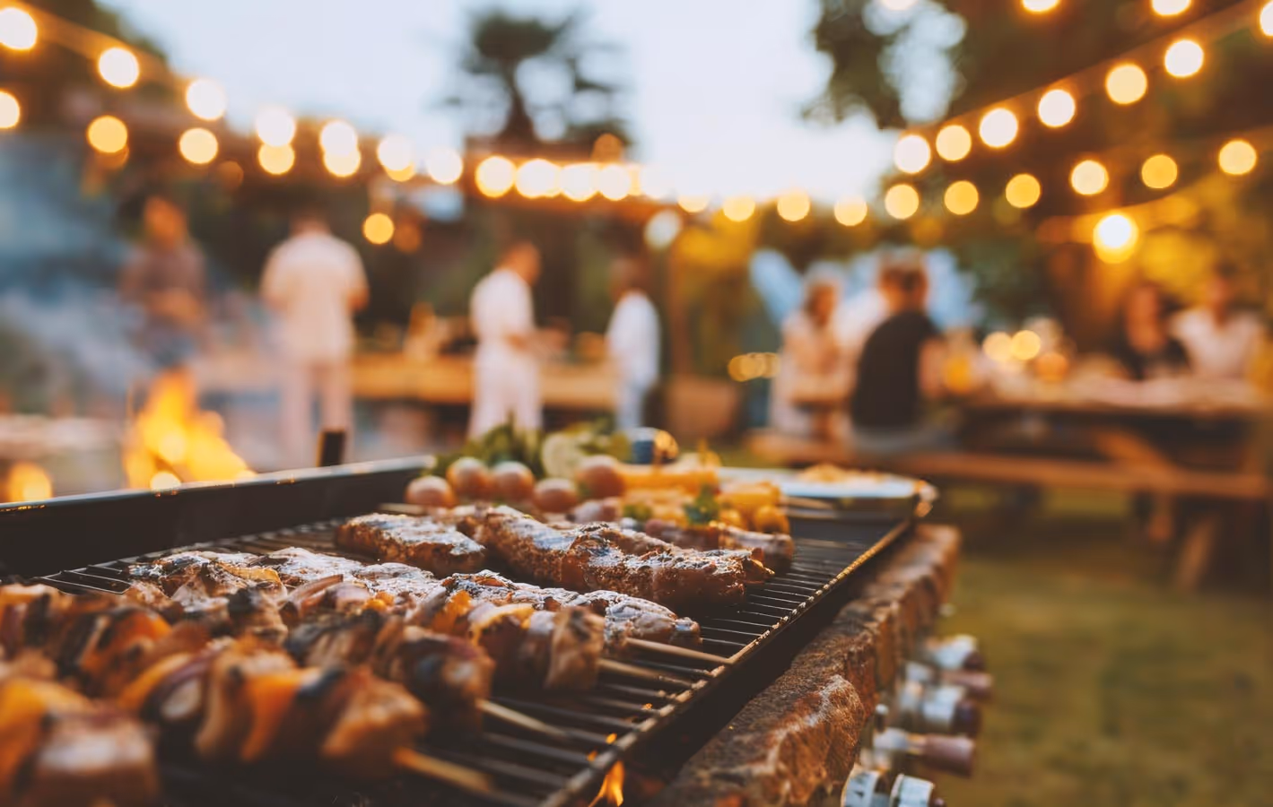 People gather outdoors for a barbecue with meats on the grill and cafe lights in the background