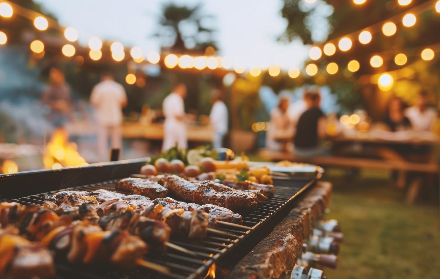 People gather outdoors for a barbecue with meats on the grill and cafe lights in the background