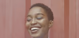 A close-up portrait of a laughing Black woman with short, natural hair against a warm, plain background.