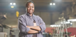 A Black person with short hair and a plaid shirt smiles while standing in a workshop setting.