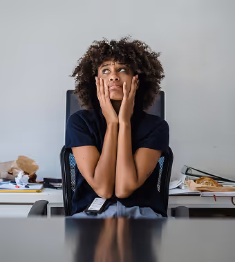 A Black woman with an afro sits at her desk with a pensive look, her hands framing her face.