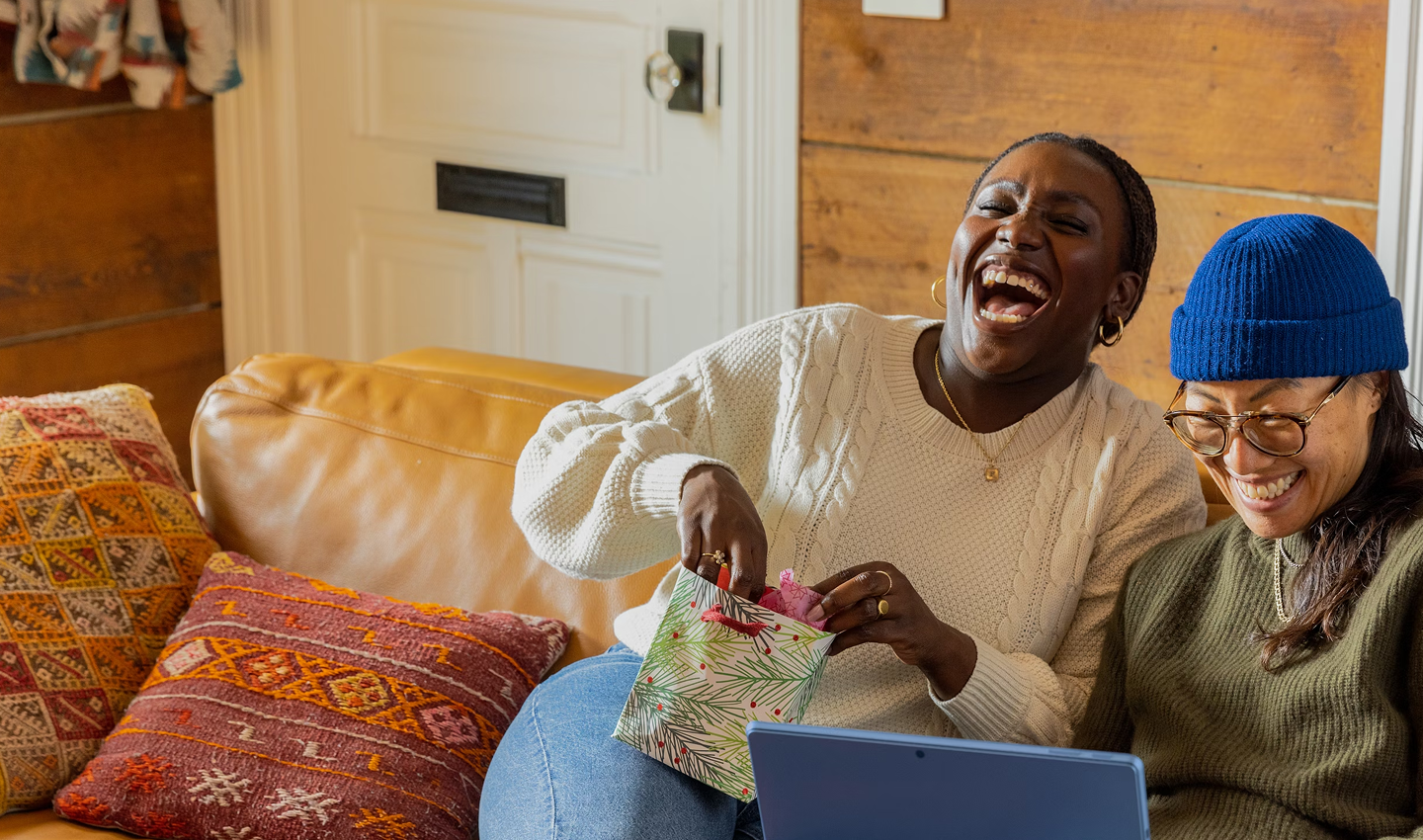 Two women sit on a couch, laughing as they stare at a laptop screen.