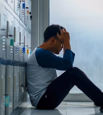 A man sits on the floor leaning against lockers, head in his hands, conveying a sense of sadness or stress.