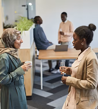 A woman wearing a hijab has a conversation with a female colleague while standing.