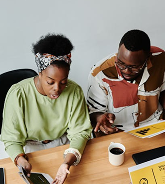 A black man and woman sit at a table staring at a screen and deliberating.