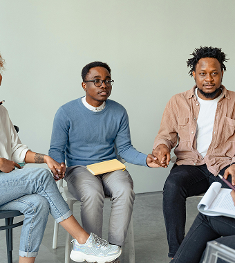 Three people sit in a circle holding hands and staring at a therapist who is out of frame.