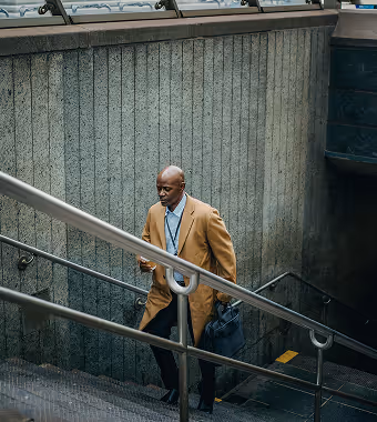 An older Black man wearing a tan coat walks up an outdoor concrete stairway.