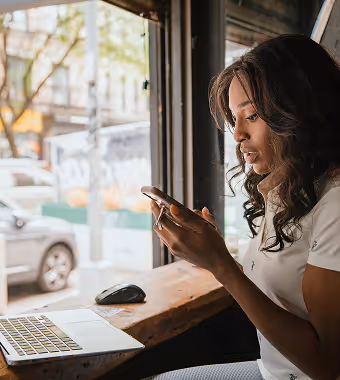 A Black woman with wavy hair stares at her phone with a laptop open in front of her seated near a Cafe window.