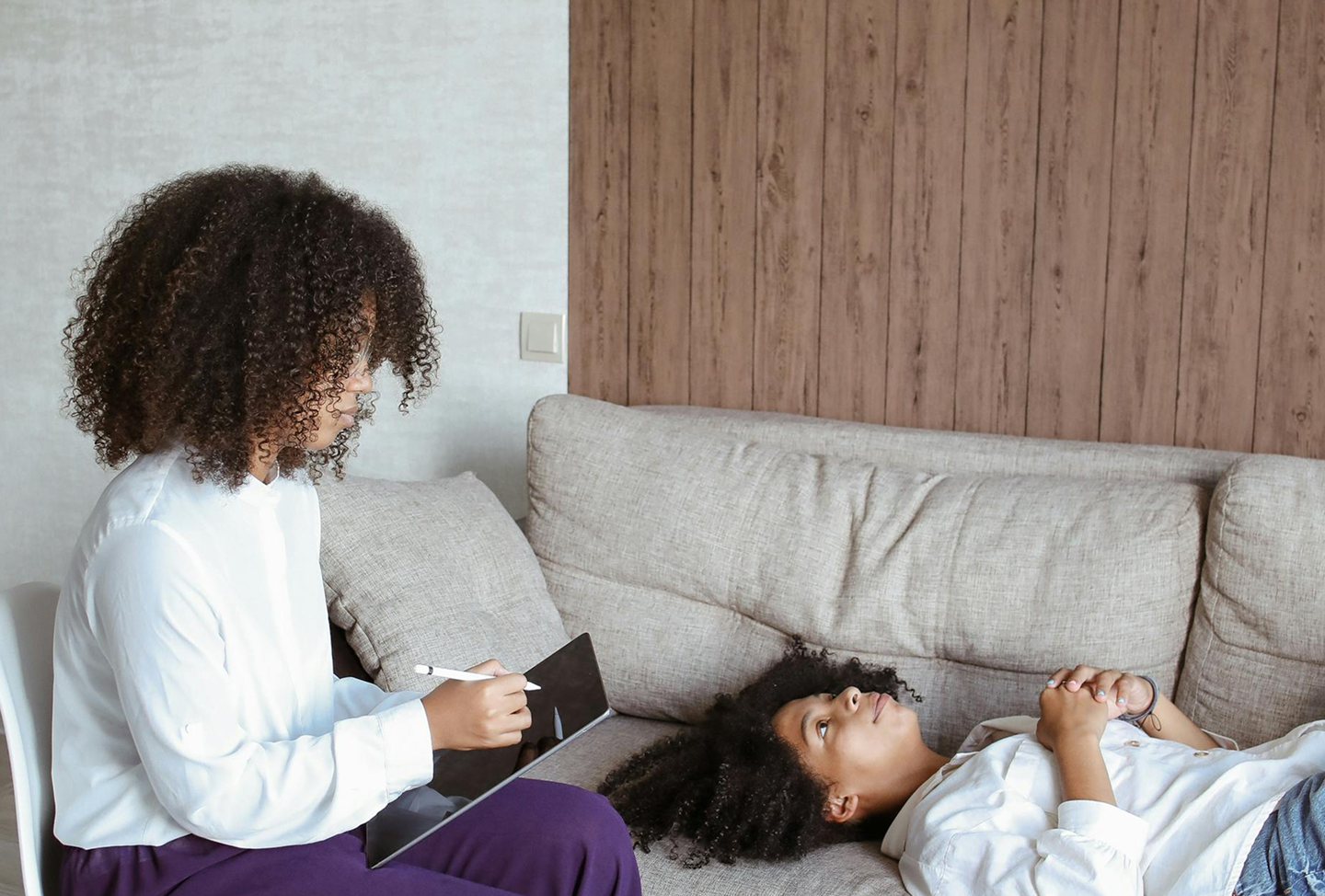 A woman with a laptop is sitting on a couch next to a girl laid on the couch.