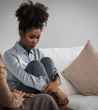 A young Black woman sits on the floor with her arms around her knees, looking down with a sad pensive expression.