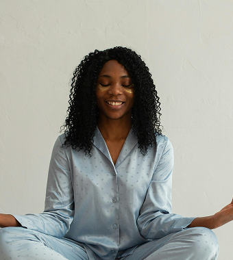 A smiling Black woman with long curly hair, wearing a silky light-blue pajama set, sits in a relaxed pose.