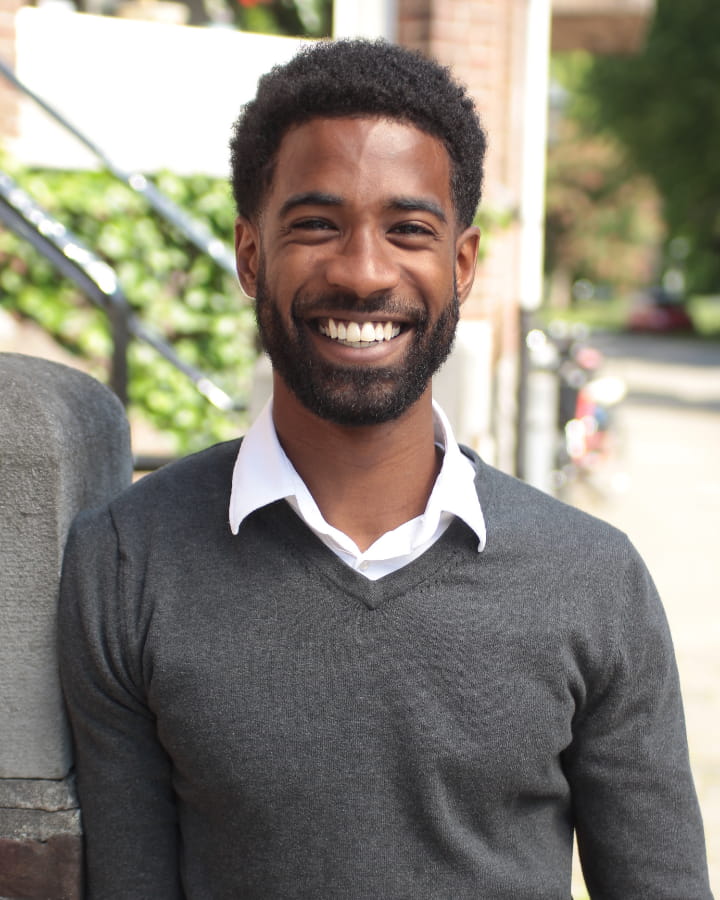 Smiling man with short curly hair and beard wearing a gray sweater over a white shirt standing outdoors.