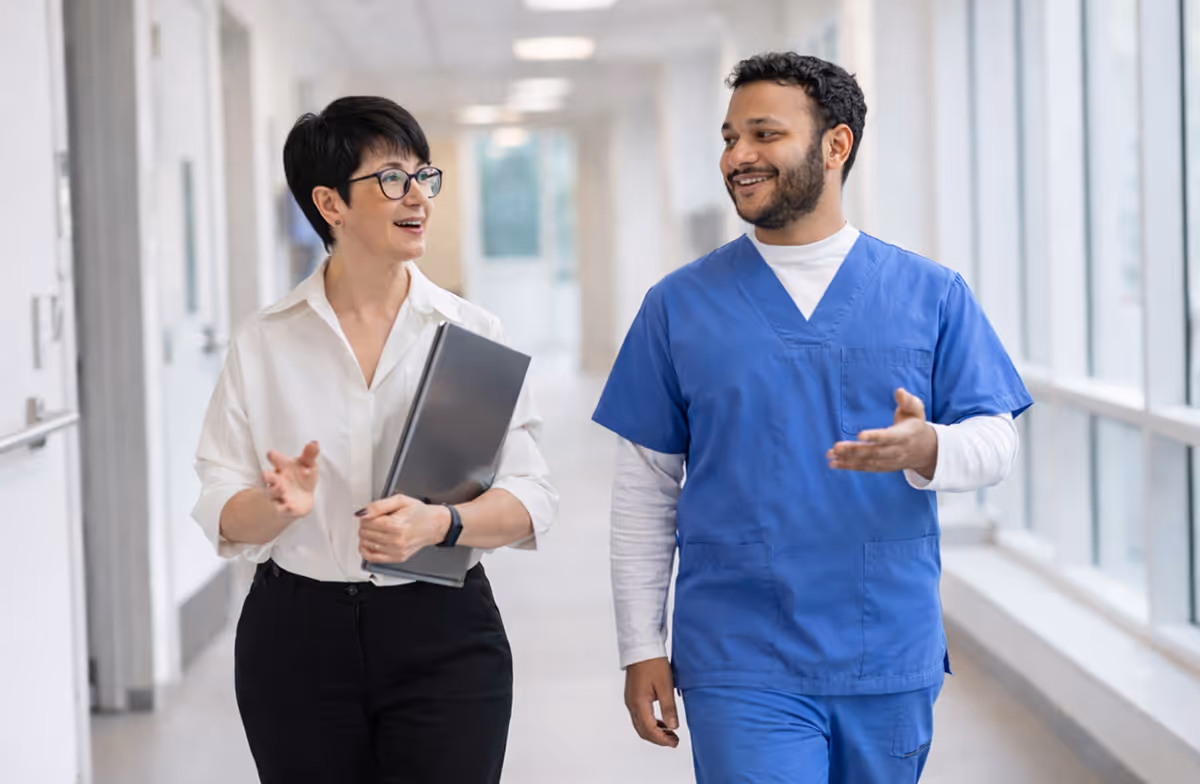 A female professional in white shirt talking with a male healthcare worker in blue scrubs while walking in a hospital corridor.