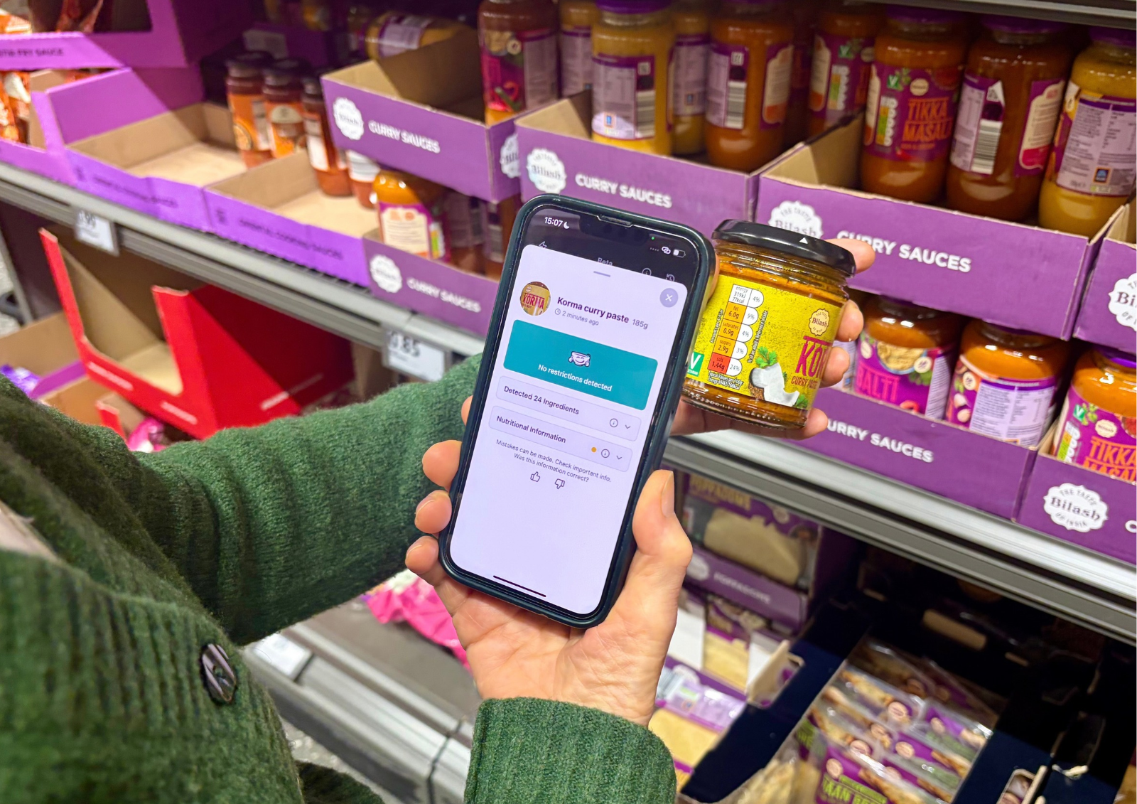 A person in a green sweater holds a jar of Korma curry paste in one hand and a smartphone in the other, displaying an app screen confirming no restrictions detected for the product, in front of shelves stocked with various curry sauces in a grocery store.