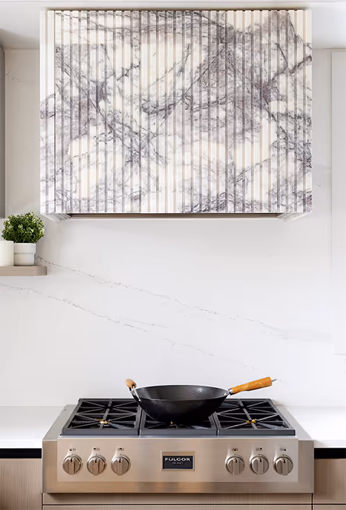 Cladded hoodfan in a flutted stone over a gas range set on a minimalist white stone countertop