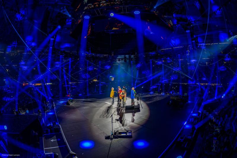 Dark circular stage with 4 men wearing primary color jackets playing instruments in teh middle of teh stage under the spotlight surrounded by multiple blue hued lights swirling around.