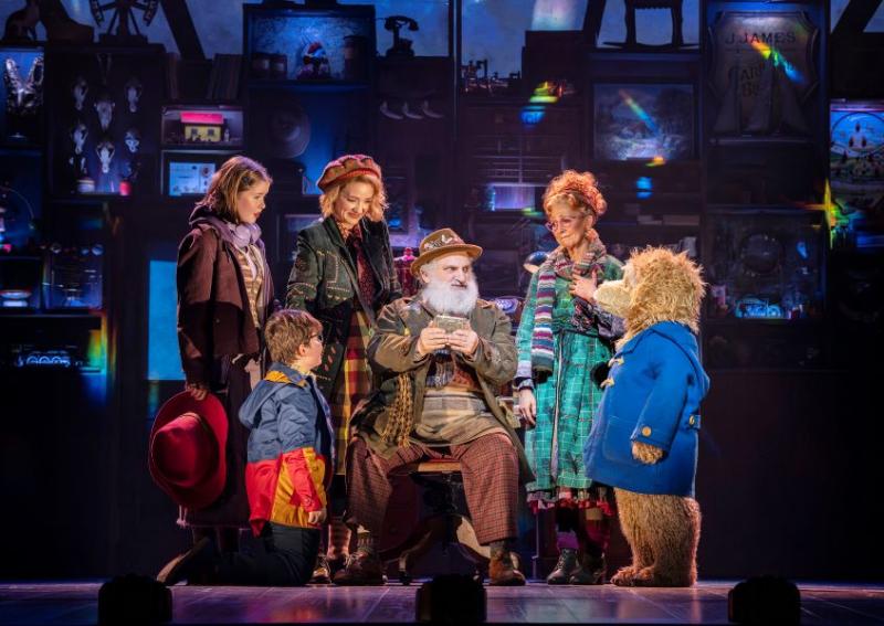 Group of scrappy indiviudlas of all ages sitting around an older man talking to a small bear wearing a cobalt blue coat.