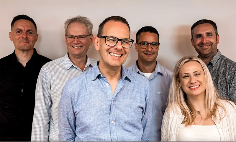 A group of smiling people with a white background, five men and a woman with long blond hair.