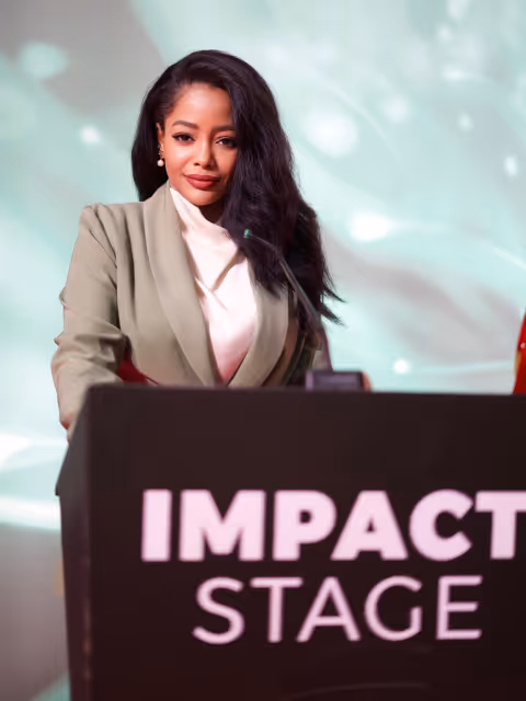 Woman with long dark hair standing behind a podium labeled 'IMPACT STAGE' with a microphone in front of her.