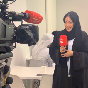 Female reporter in a black abaya and headscarf holding a red Al Arabiya microphone in front of a video camera in a white indoor setting.