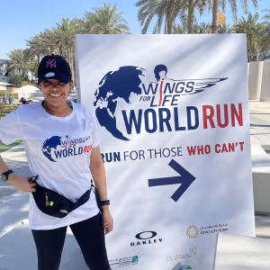 Smiling woman in a white Wings for Life World Run T-shirt and black cap standing next to a Wings for Life World Run sign with event logos and an arrow pointing right.