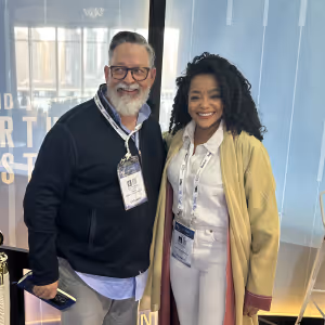 Smiling older man with glasses and a badge standing next to a smiling young woman with long curly hair, wearing a light yellow jacket and white outfit.