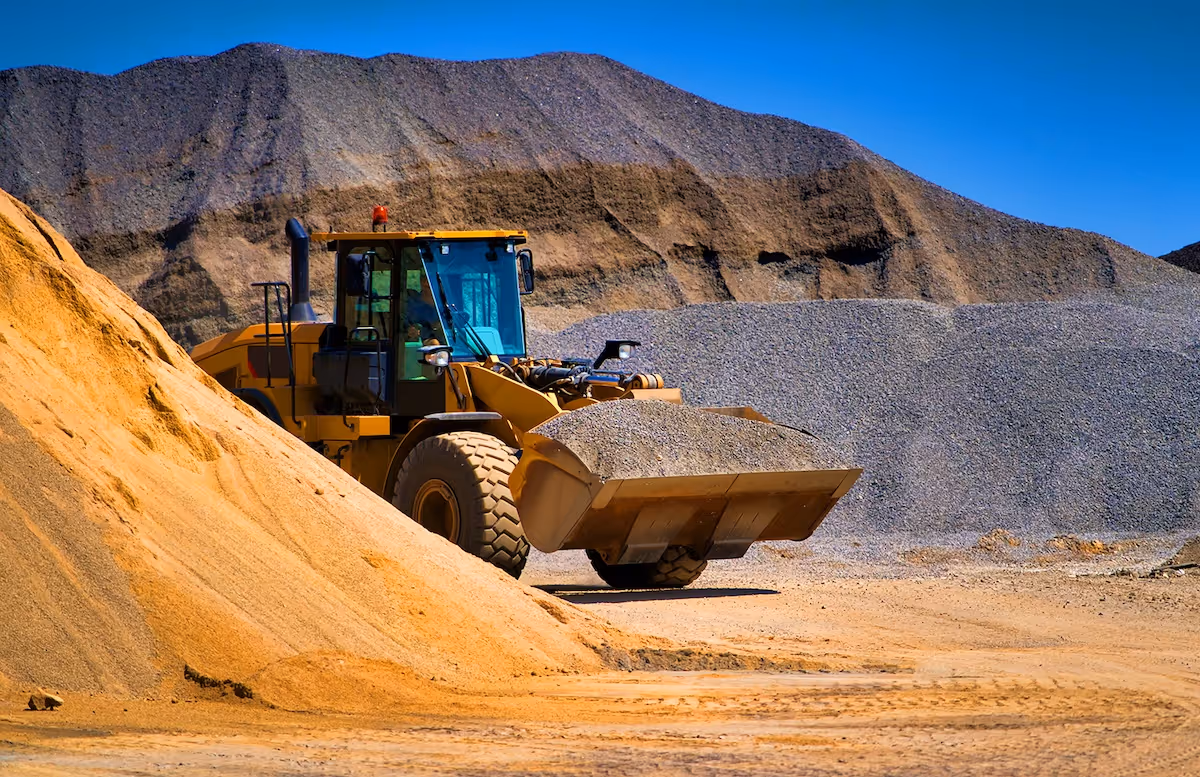 Yellow front loader carrying gravel in a quarry with large sand and gravel piles can see extended life with specialty lubricants