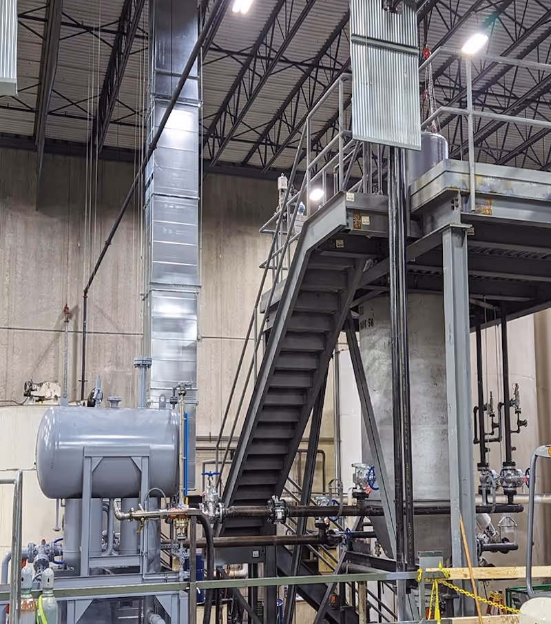 Industrial lubricant plant interior with metal staircase, large tanks, pipes, and ventilation ducts under high ceiling.