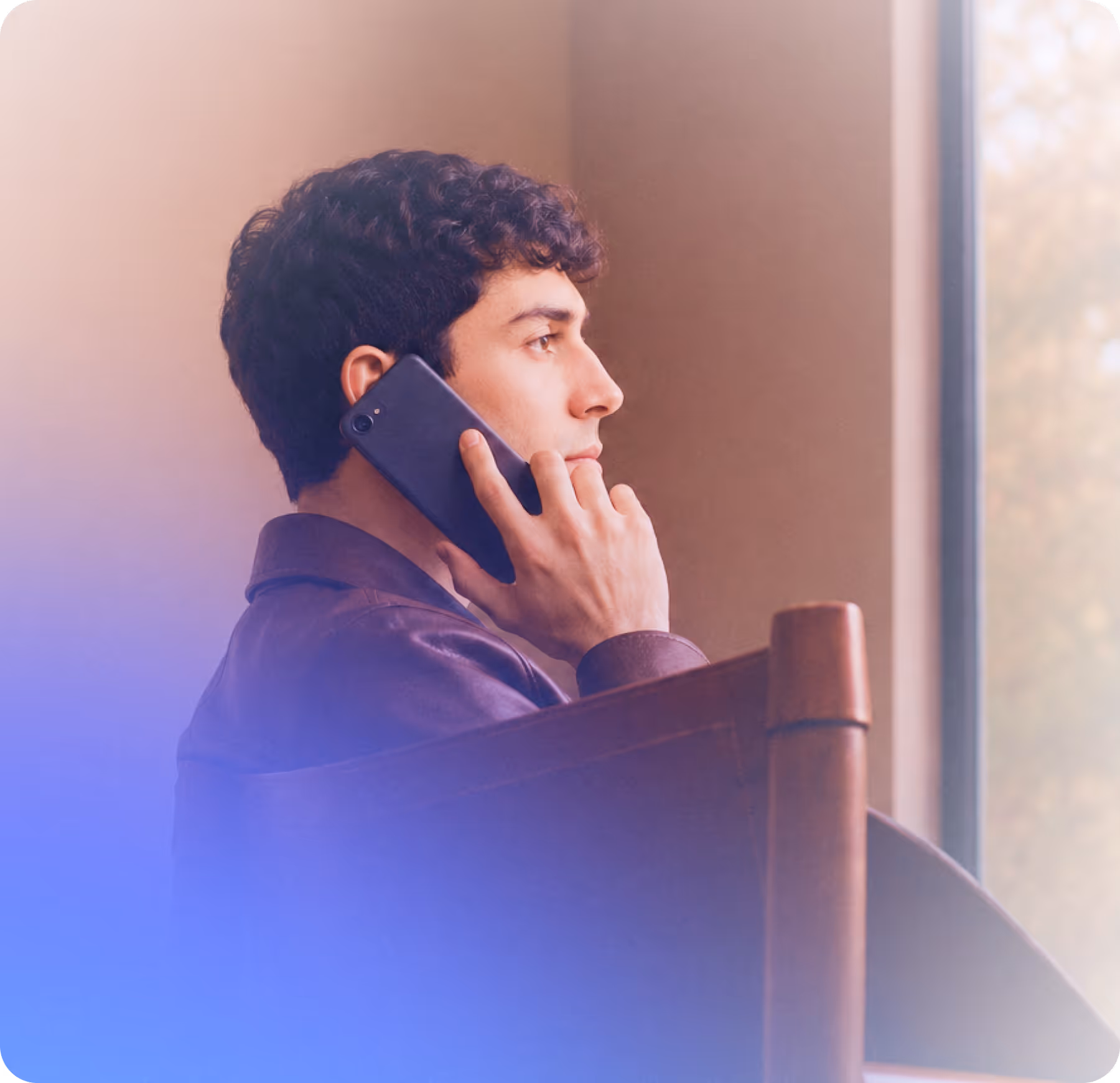 Young man with curly hair sitting in a chair, talking on a smartphone near a window.