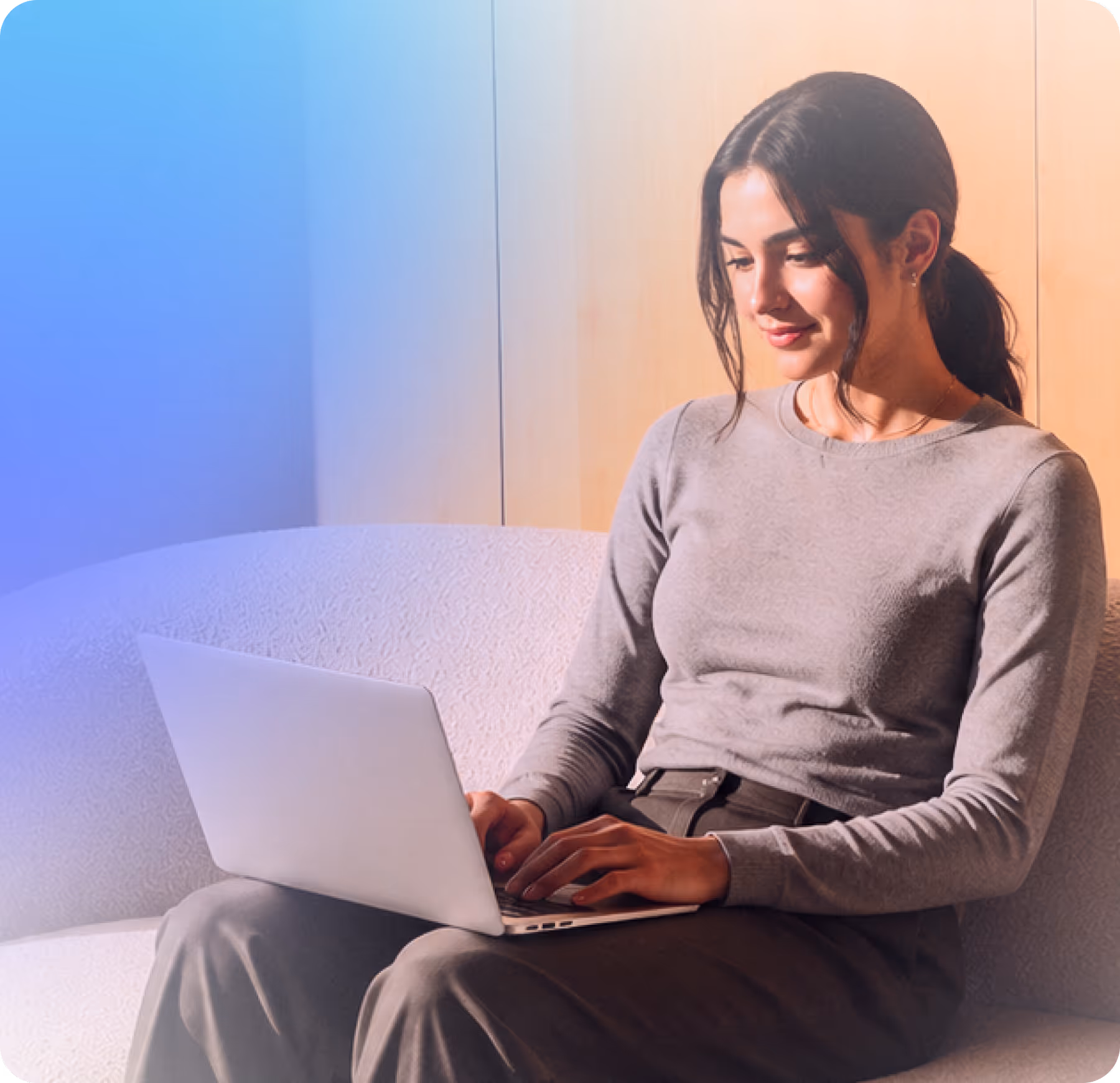 Young woman sitting on couch typing on a laptop with a soft smile on her face.