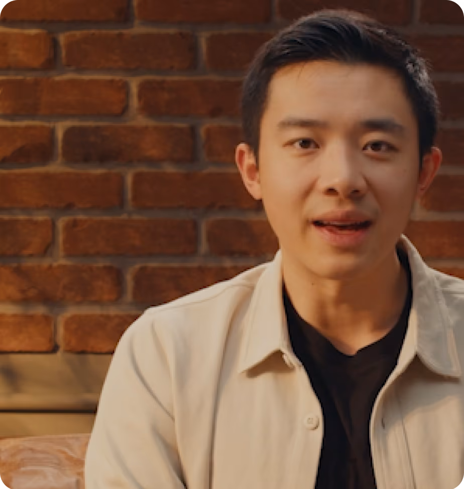 Young man in a light jacket speaking, with a brick wall in the background.