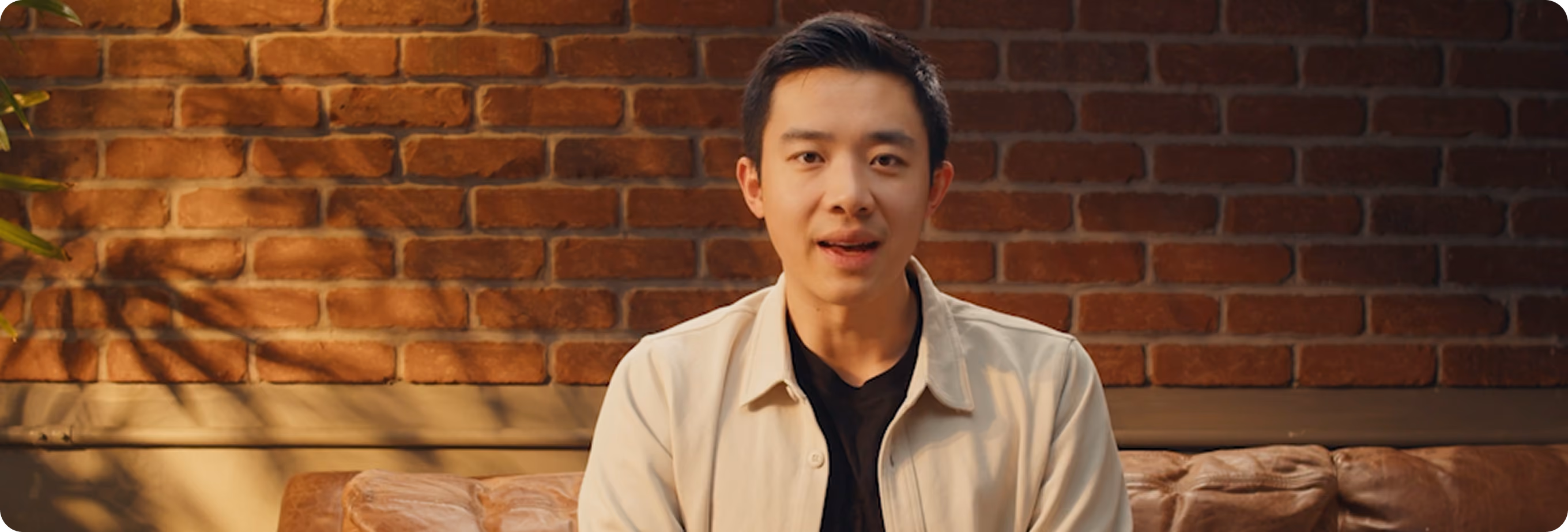 Young man with black hair sitting on a brown leather couch against a brick wall background.
