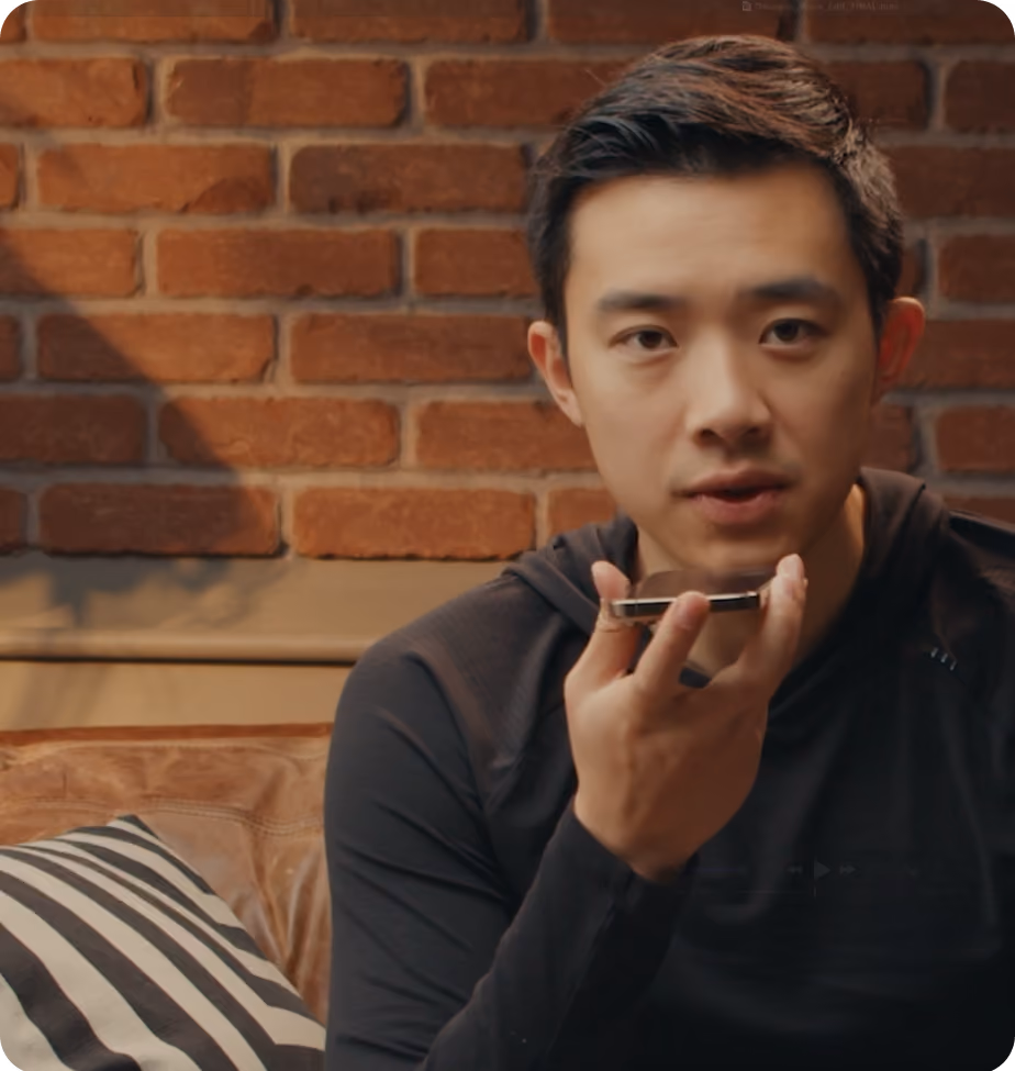 Man in black shirt speaking into smartphone held near his mouth, seated on couch with brick wall behind.