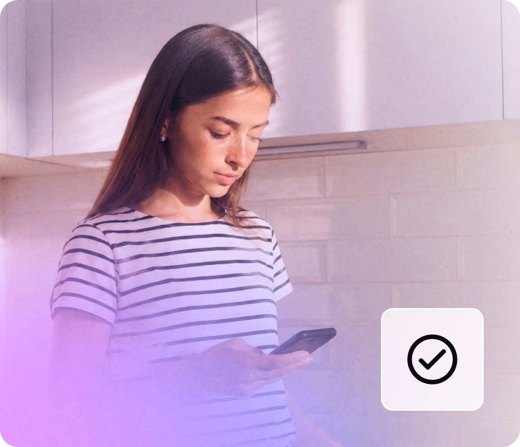 Young woman in striped shirt looking at her smartphone in a kitchen with white cabinets.