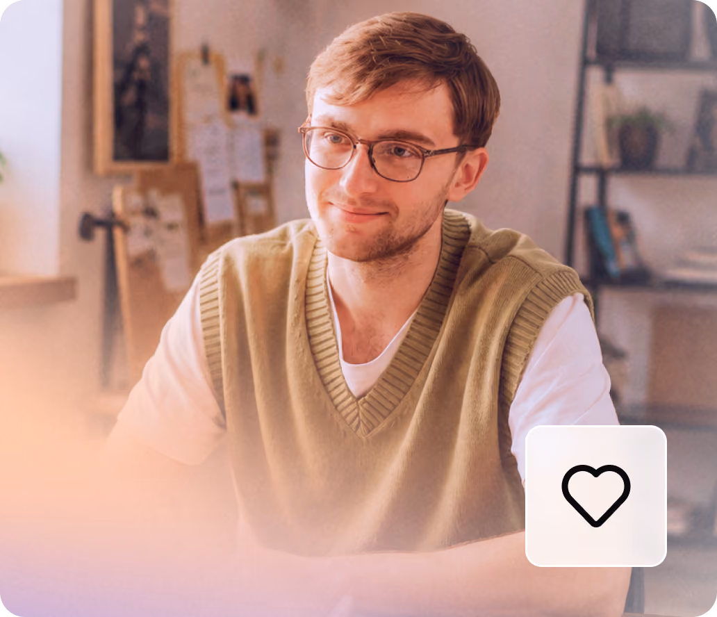 Smiling young man with glasses and a green vest sitting at a desk in a softly lit room.