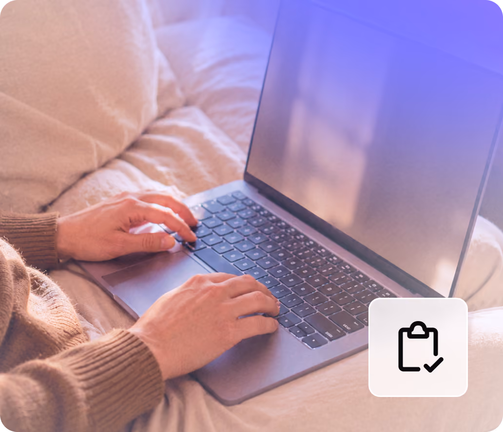 Person typing on a laptop keyboard while sitting on a bed with beige bedding.
