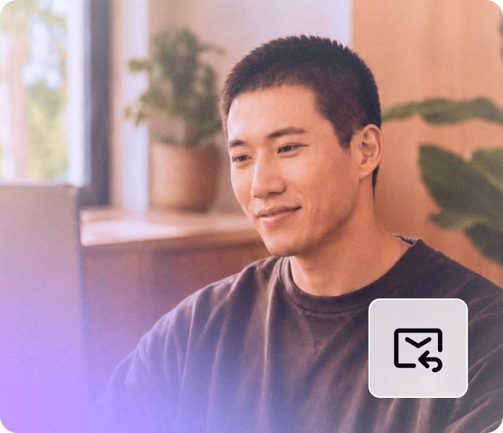 Young man with short hair smiling and looking at a laptop in a cozy indoor setting with plants.