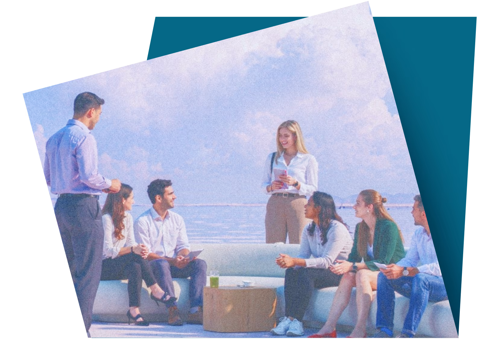 Group of seven casually dressed people talking outdoors on white bench with ocean in background.