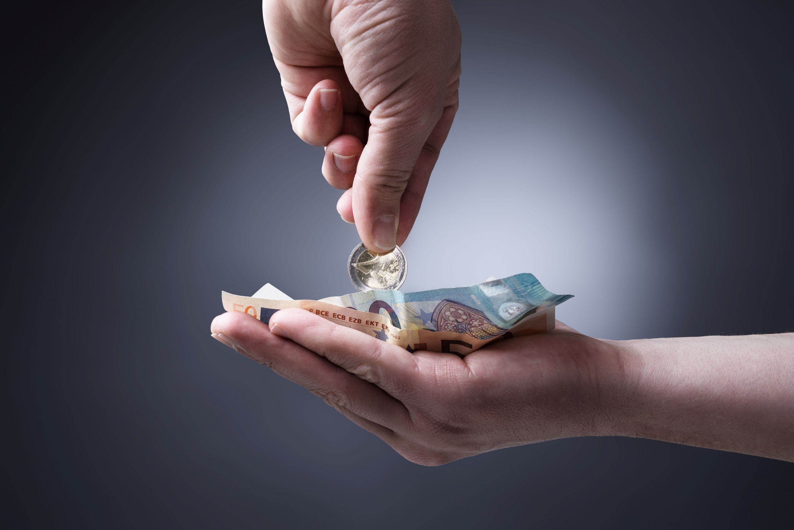 Eine Hand hält gefaltete 50-Euro-Scheine, während eine andere Hand eine Euro-Münze darüber hält.