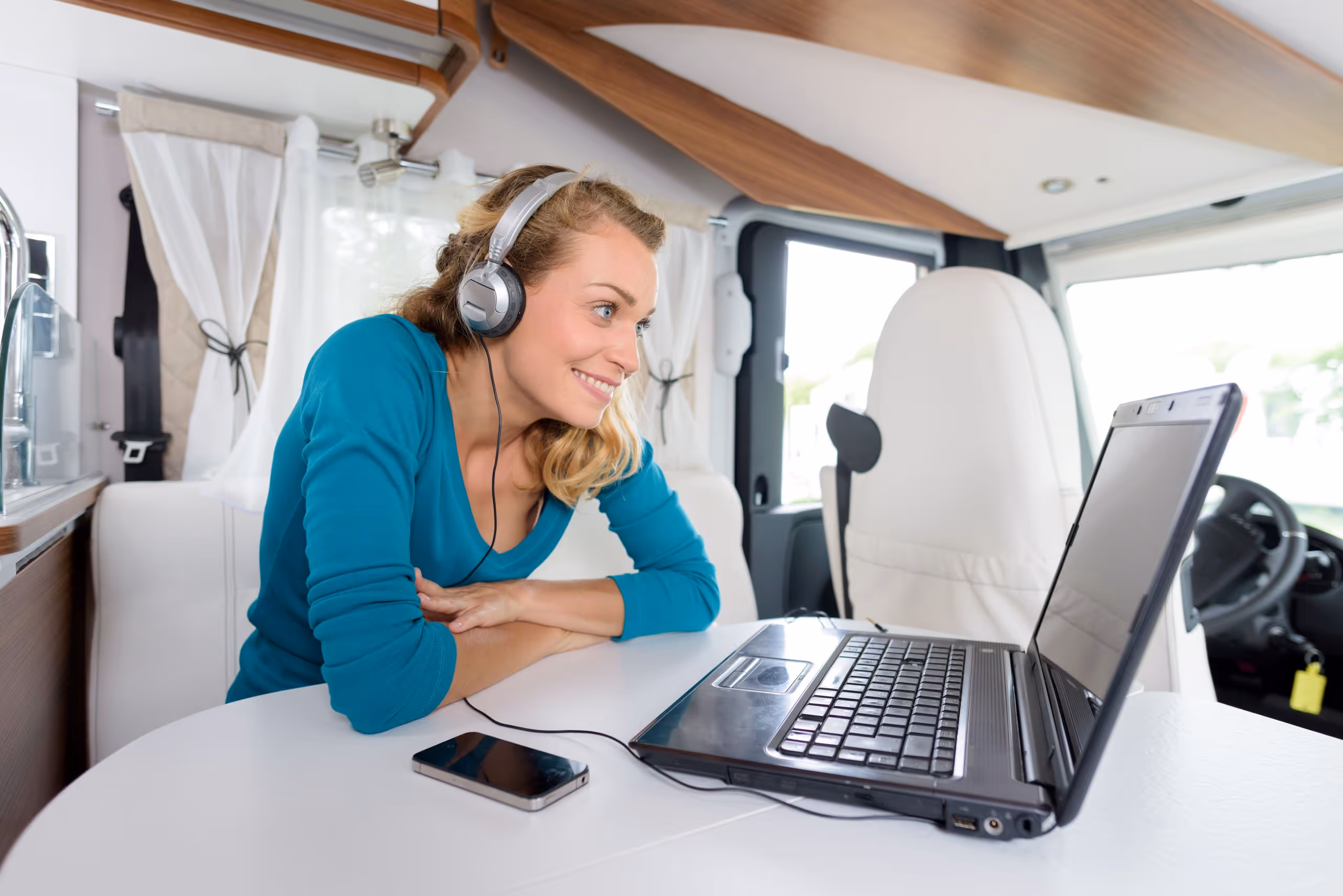 Frau mit Kopfhörern sitzt in einem Wohnmobil und schaut lächelnd auf einen Laptop auf dem Tisch.