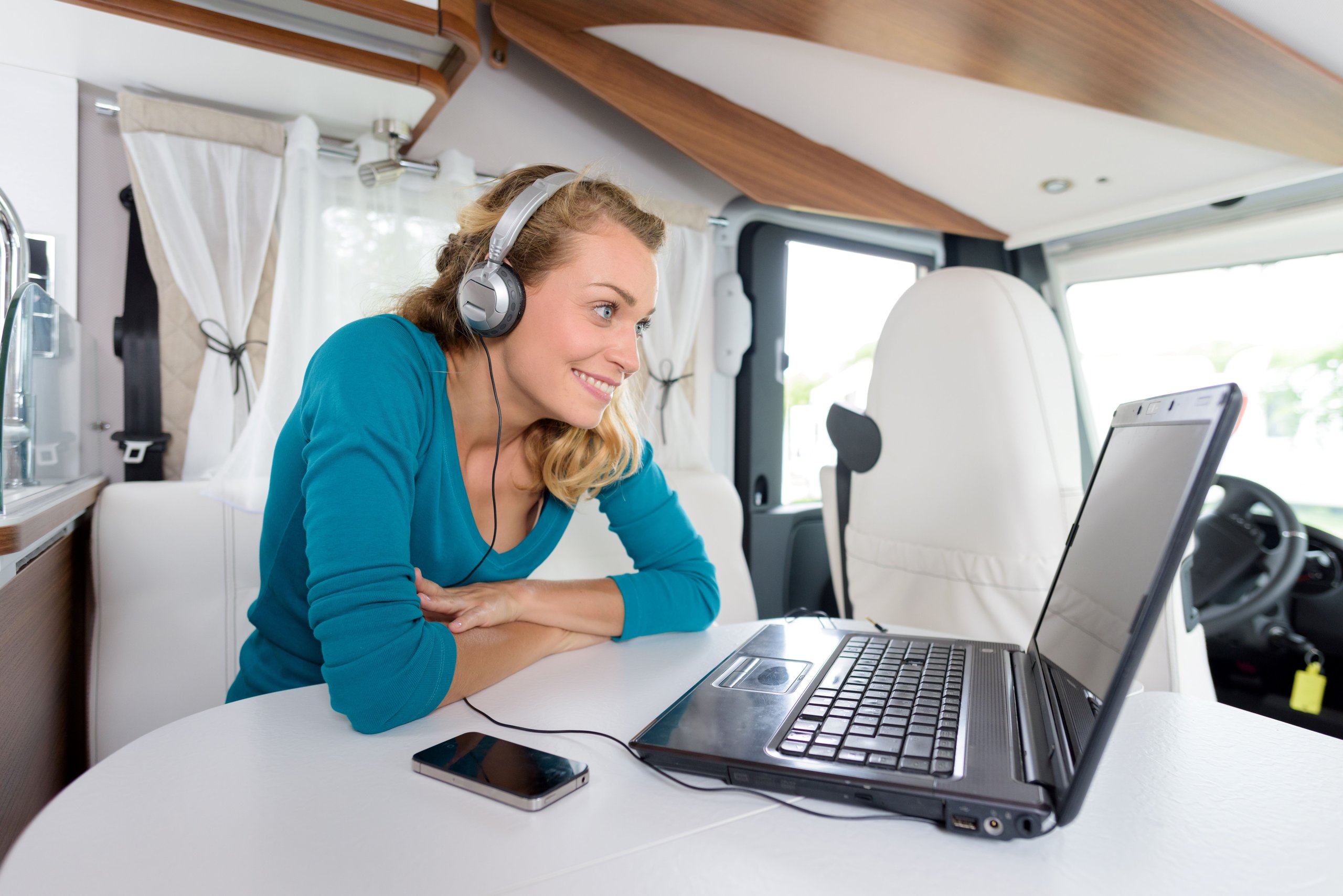 Frau mit Kopfhörern sitzt in einem Wohnmobil und schaut lächelnd auf einen Laptop auf dem Tisch.